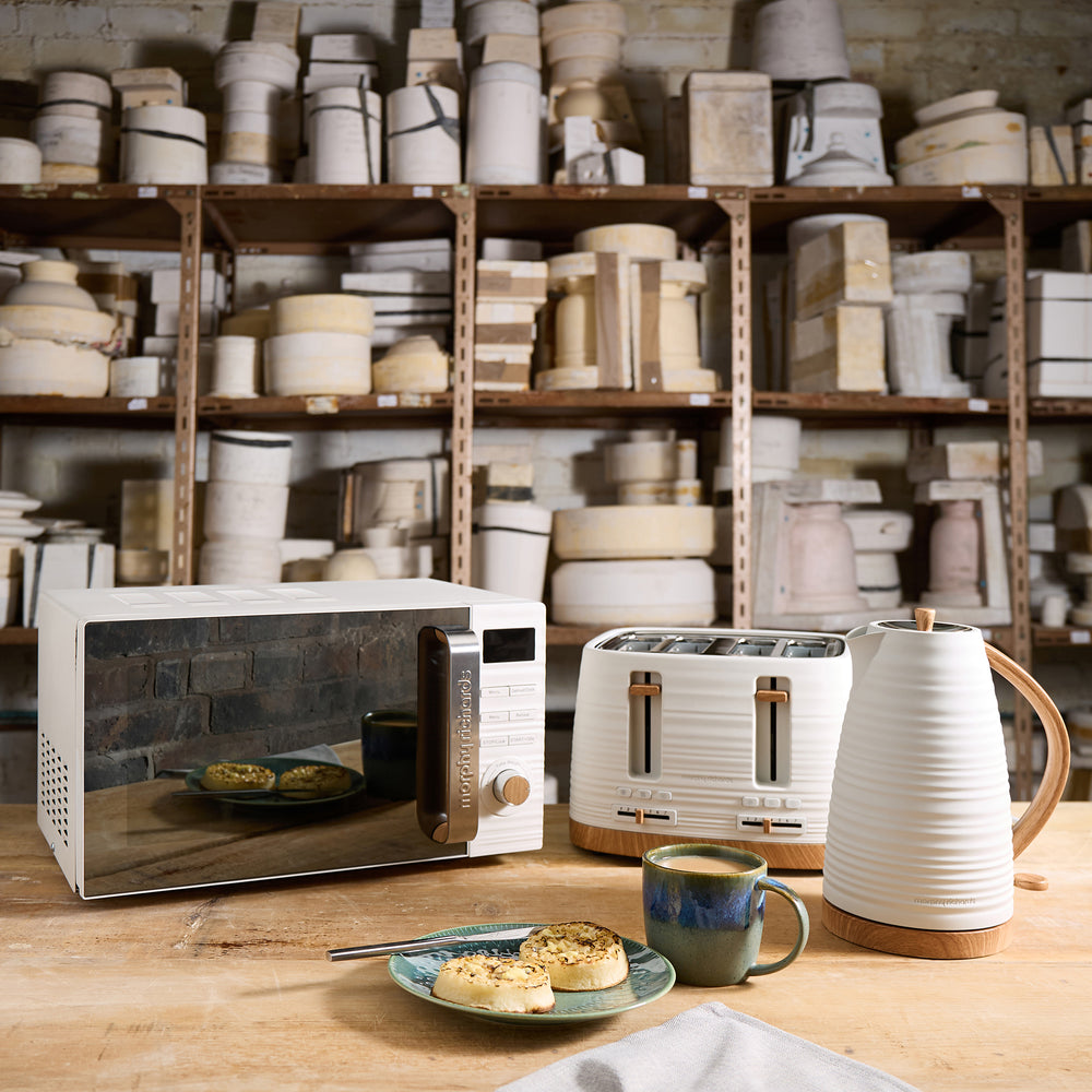 Kitchen appliances including a microwave, toaster, and kettle on a wooden table with shelves of kitchenware in the background.